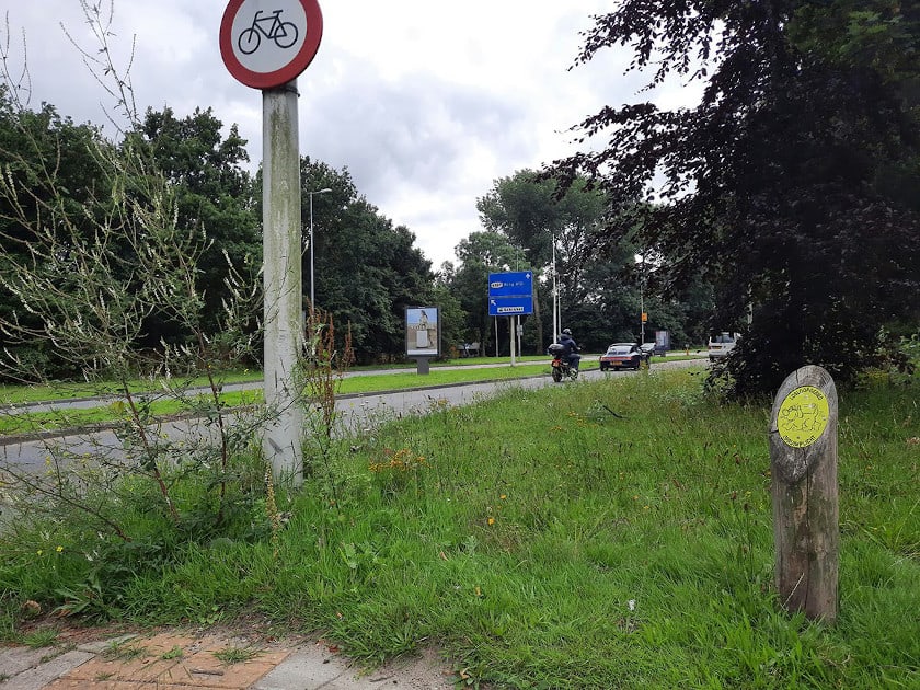 Een doorgaande weg met daarnaast een groenstrook waar een verbodsbord voor fietsers staat, die niet op de doorgaande weg mogen komen, met ongeveer een meter daarnaast een paaltje met een teken dat hier een hondenlosloopgebied begint.